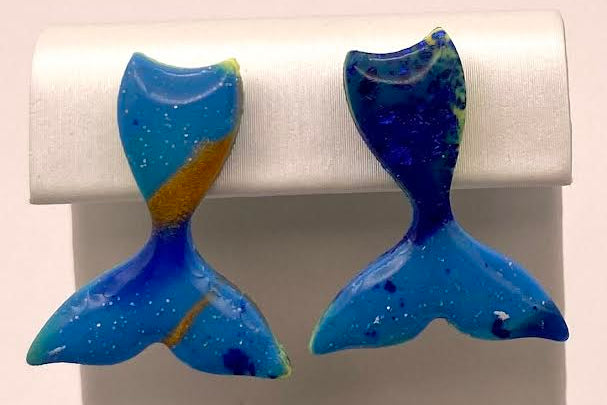 A pair of handmade blue and yellow mermaid tail stud earrings displayed on a white plastic jewelry stand against a light pink background.