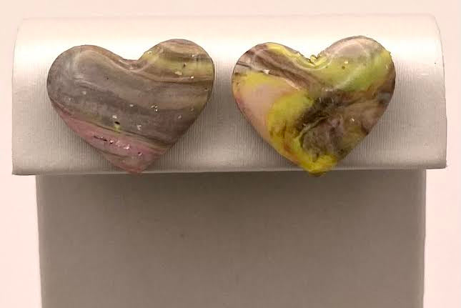 Two heart-shaped marbled resin cabochons, one with pink, gray, and gold swirls and the other with yellow, green, and brown swirls, displayed on a white jewelry stand.