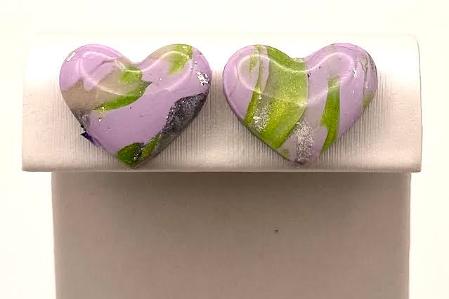 A pair of heart-shaped glass stud earrings with a marbled pattern of lavender purple, lime green, and gray, displayed on a white rectangular stand against a white background.