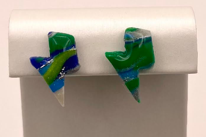 A pair of handmade lightning-bolt stud earrings made from marbled green, blue, and white glass, displayed on a white jewelry stand.