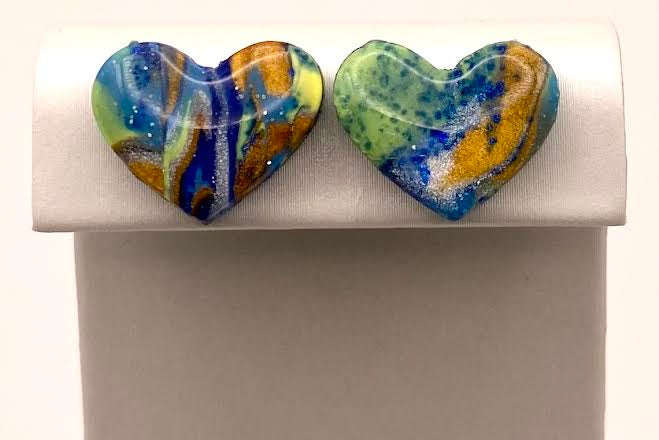 A pair of heart-shaped glass stud earrings with a marbled swirl pattern of blue, green, yellow, and white, displayed on a white rectangular jewelry stand.