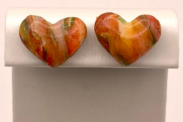 A pair of heart-shaped marbled gemstone stud earrings in orange, red, and green, displayed on a white jewelry stand.