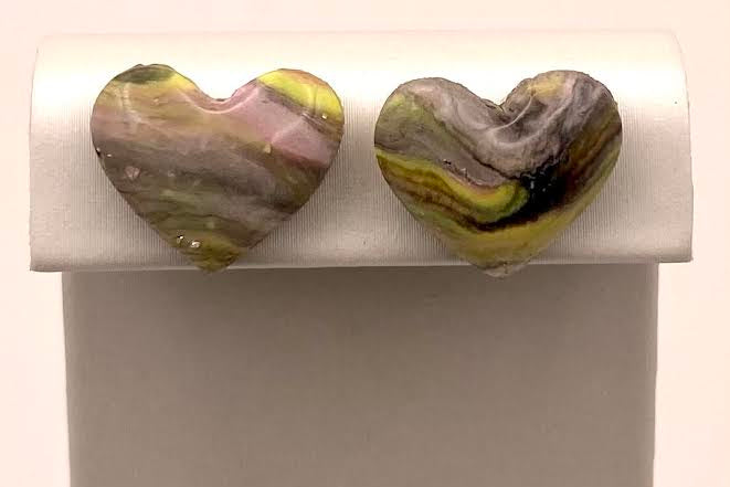 A pair of heart-shaped stud earrings with marbled, stone-like patterns in olive green, yellow, pink, and gray, displayed on a white jewelry stand against a light background.