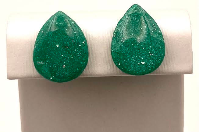 A pair of teardrop-shaped green glitter stud earrings displayed on a white jewelry stand against a white background.