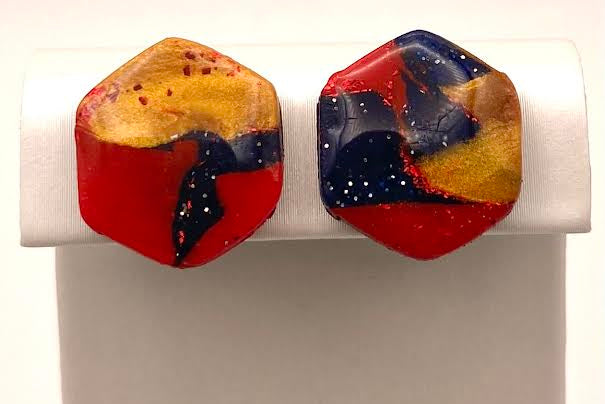 A pair of hexagonal stud earrings with a marbled abstract design in red, yellow, and navy blue, displayed on a white jewelry stand against a white background.