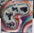 A square resin art piece featuring a Geode design made from two interlocking geode-like crescents filled with clear resin and embedded dark and purple crystals, set against a marbled background of orange, purple, and blue swirls.