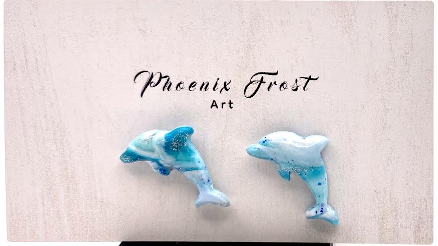 A pair of dolphin-shaped earrings in light blue and white with darker blue speckles, displayed on a light pink card with the brand name 'Phoenix Frost Art' printed above them.