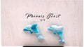 A pair of dolphin-shaped earrings in light blue and white with darker blue speckles, displayed on a light pink card with the brand name 'Phoenix Frost Art' printed above them.