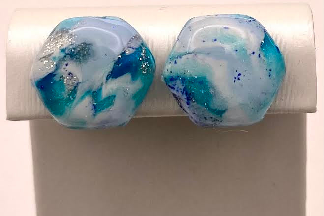 A pair of hexagonal stud earrings with a marbled blue, white, and teal glass design, displayed on a white jewelry stand against a light background.