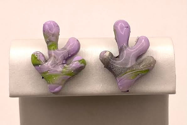 A pair of small coral-shaped stud earrings in lavender purple and green, displayed on a white jewelry stand against a light pink background.