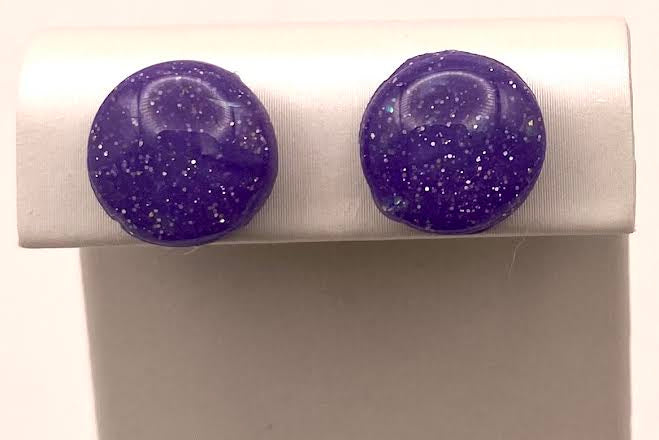 A pair of round stud earrings with a deep purple glitter finish, displayed on a white jewelry stand against a white background.