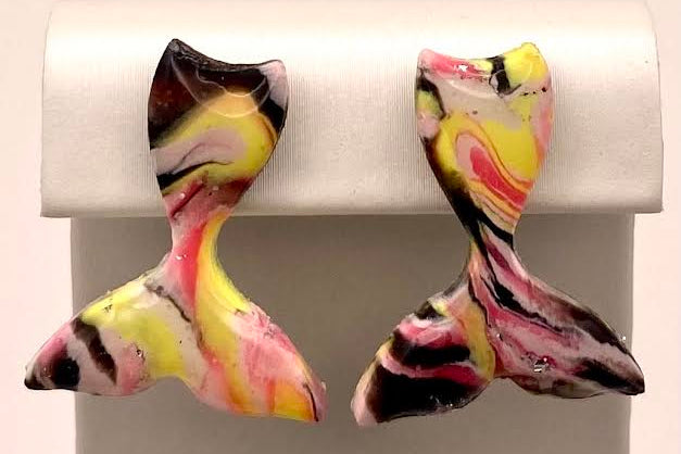 A pair of handmade polymer clay earrings shaped like mermaid tails, featuring a marbled pattern of pink, yellow, black, and white, displayed on a white jewelry stand.