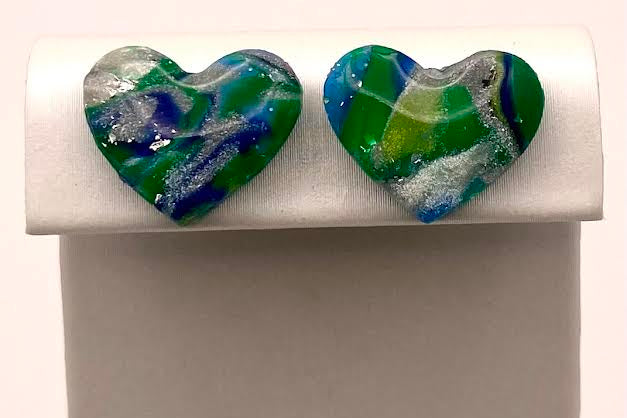 A pair of heart-shaped glass stud earrings with a marbled swirl pattern of green, blue, and white, displayed on a white jewelry stand.