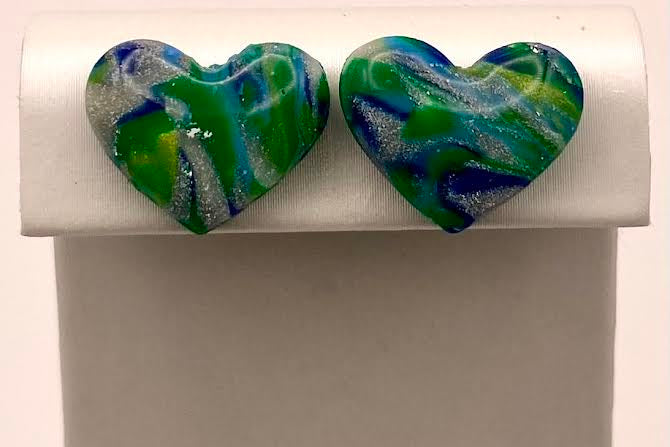 A pair of heart-shaped stud earrings with a marbled green, blue, and white pattern, displayed on a white rectangular jewelry stand against a plain white background.