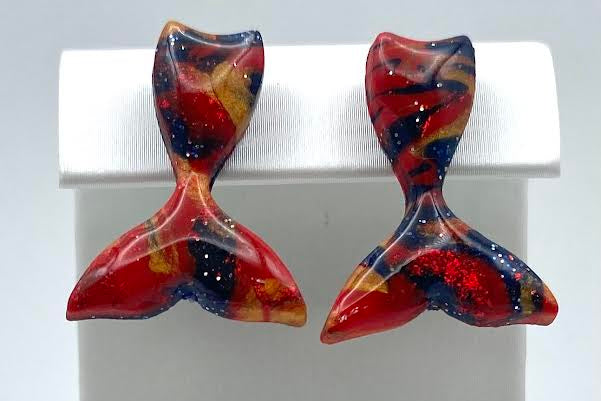 A pair of handmade mermaid tail stud earrings with a marbled red, blue, and orange pattern, displayed on a white jewelry stand.