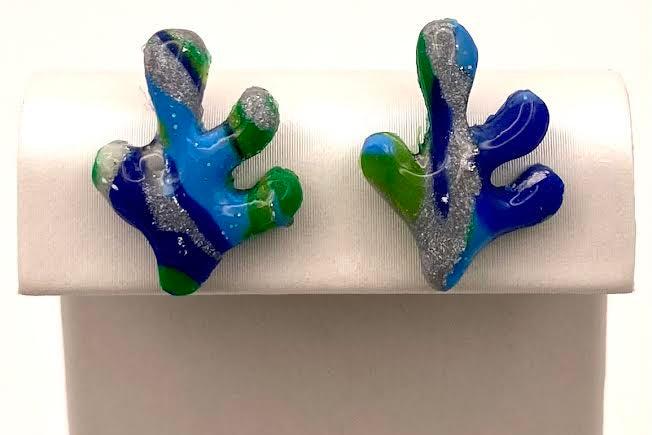 A pair of handmade coral-shaped stud earrings with blue, green, and silver glitter swirls, displayed on a white rectangular stand.