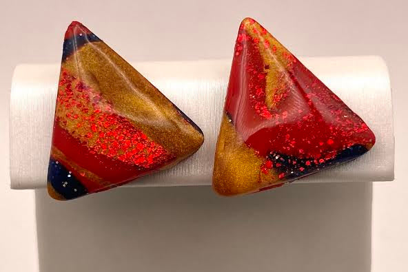 A pair of triangular stud earrings with a marbled pattern of red, orange, yellow, and dark blue, displayed on a white jewelry stand.