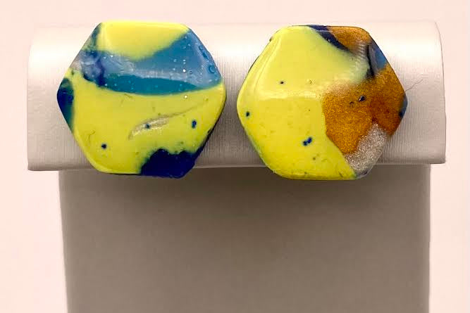 A pair of hexagonal stud earrings with a marbled pattern of bright yellow, blue, and brown, displayed on a white stand against a light pink background.