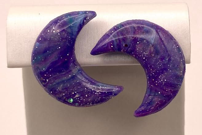 A pair of crescent moon-shaped earrings with a purple and blue galaxy glitter design, displayed on a white jewelry stand.