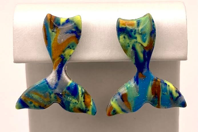 A pair of mermaid tail stud earrings with a marbled pattern of blue, green, yellow, and orange, displayed on a white jewelry stand.