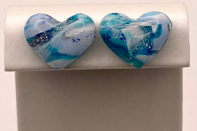 A pair of heart-shaped glass stud earrings with a marbled blue, white, and teal pattern and small glitter flecks, displayed on a white rectangular jewelry stand against a light background.