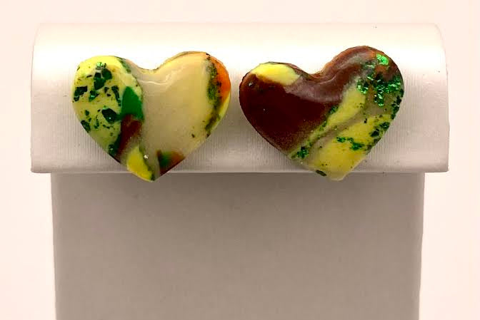A pair of heart-shaped stud earrings displayed on a white rectangular stand against a plain white background. One earring is marbled cream, green, and orange, and the other is marbled dark red, yellow, and green.