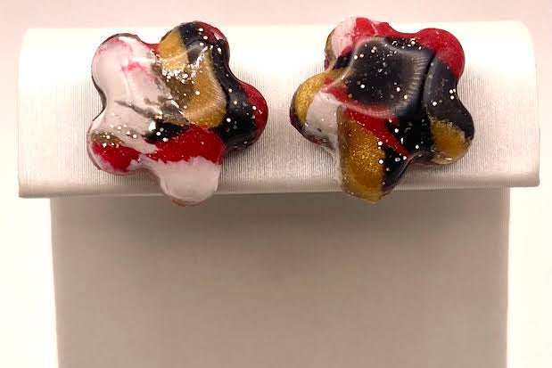 A pair of handmade stud earrings with a marbled swirl pattern of red, black, white, and gold, displayed on a white jewelry stand.