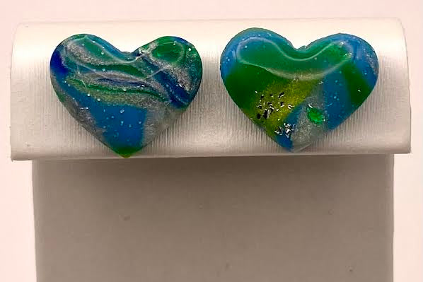 A pair of heart-shaped glass stud earrings with blue, green, and white marbled swirls, displayed on a white rectangular stand against a light background.