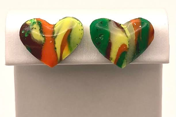 A pair of heart-shaped marbled resin stud earrings in green, yellow, orange, and brown, displayed on a white rectangular stand against a white background.
