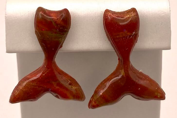 A pair of small fish-tail shaped stud earrings in a glossy reddish-brown resin with lighter cream highlights, displayed on a white plastic jewelry stand.