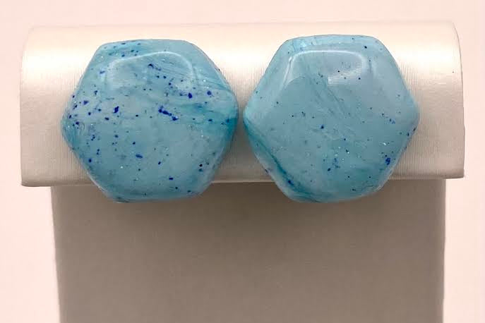 A pair of faceted light blue hexagonal stud earrings with dark blue speckles, displayed on a white jewelry stand against a light background.