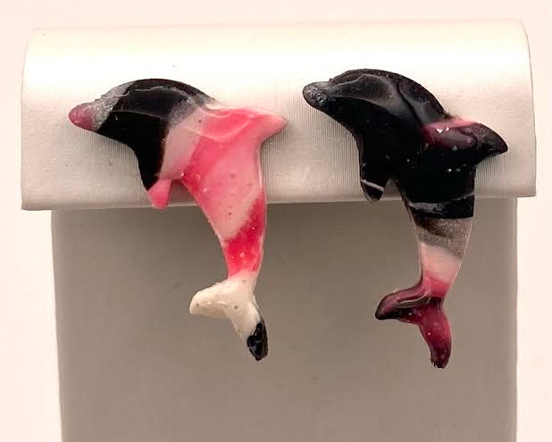 A pair of small dolphin-shaped earrings displayed on a white jewelry stand against a light background. The dolphins have black, pink, white, and dark red marbled patterns.
