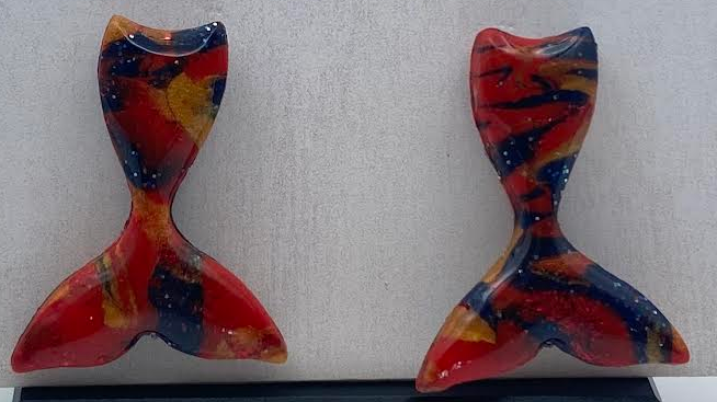 A pair of handmade resin earrings shaped like mermaid tails, featuring a marbled pattern of red, orange, yellow, and dark blue, displayed on a white textured card with the brand name 'Phoenix Frest Art'.