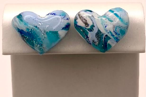 A pair of heart-shaped glass stud earrings with a marbled blue, white, and teal pattern, displayed on a white jewelry stand.
