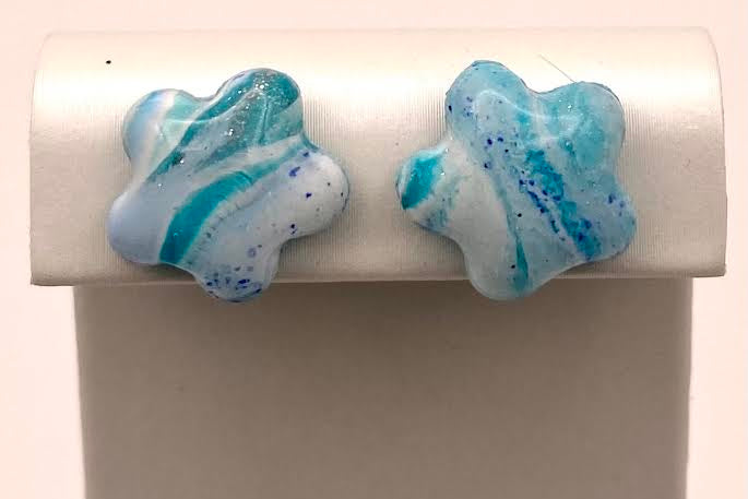 A pair of handmade star-shaped stud earrings with a marbled blue, white, and light teal glass finish, displayed on a white rectangular jewelry stand.