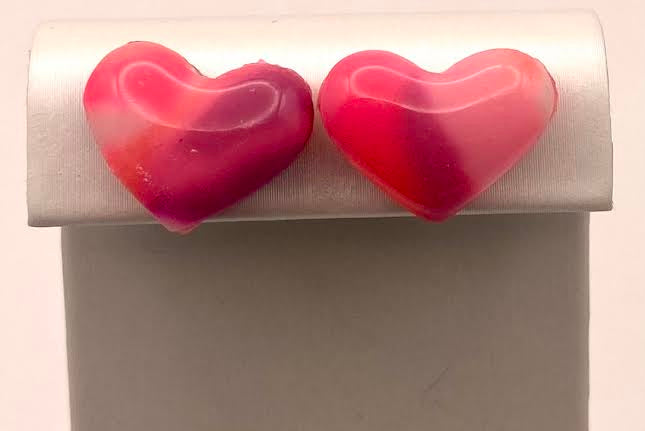 A pair of heart-shaped stud earrings in a pink and red marbled pattern, displayed on a white jewelry stand against a light background.