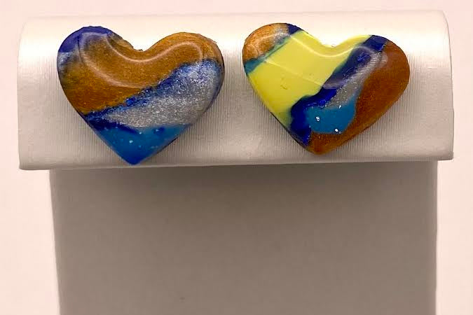 A pair of heart-shaped marbled glass stud earrings displayed on a white jewelry stand. The earrings feature swirls of blue, yellow, orange, and white.