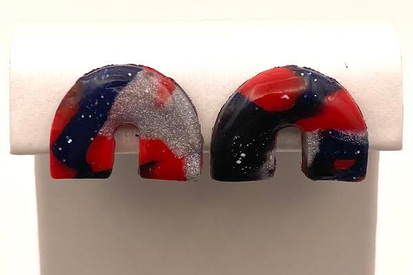 A pair of small U-shaped earrings with a marbled pattern of red, navy blue, and glittery silver, displayed on a white stand.