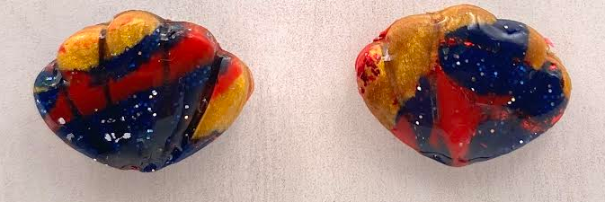 A pair of handmade, irregularly shaped clay or glass art pieces in a marbled swirl of red, yellow, and dark blue, displayed on a light beige wooden sign with the text 'Phoenix Frost Art'.