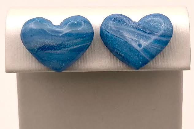 A pair of heart-shaped blue marbled stud earrings displayed on a white jewelry stand against a light background.