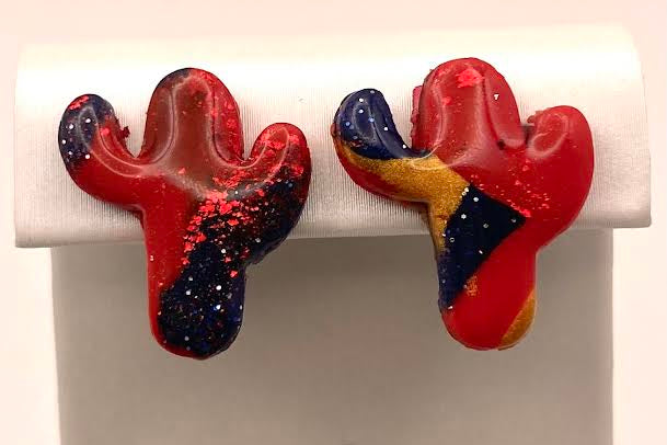A pair of cactus-shaped stud earrings with a glossy red, navy blue, and yellow marbled pattern, displayed on a white jewelry stand.
