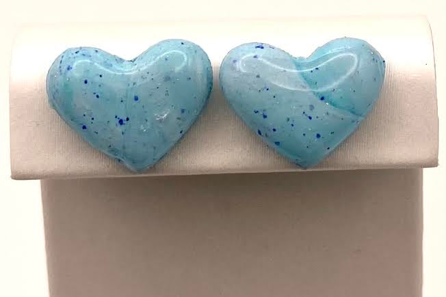 A pair of light blue heart-shaped stud earrings with dark blue speckles, displayed on a white jewelry stand against a white background.