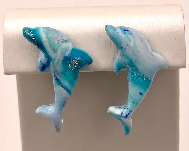 A pair of dolphin-shaped stud earrings in a marbled blue, white, and turquoise glaze with small silver glitter flecks, displayed on a white jewelry stand.
