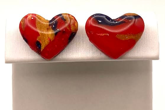 A pair of heart-shaped glass stud earrings with a marbled red, orange, and black pattern, displayed on a white jewelry stand against a white background.