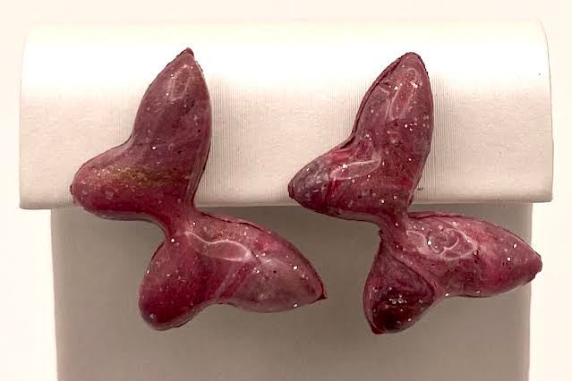 A pair of small, butterfly-shaped stud earrings in a glossy, deep magenta-pink resin with tiny glittery flecks, displayed on a white plastic jewelry stand.
