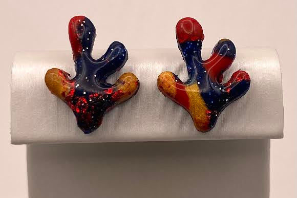 A pair of handmade glass stud earrings with a coral or antler-like shape, featuring a blue base with red, orange, and yellow accents, displayed on a white jewelry stand.
