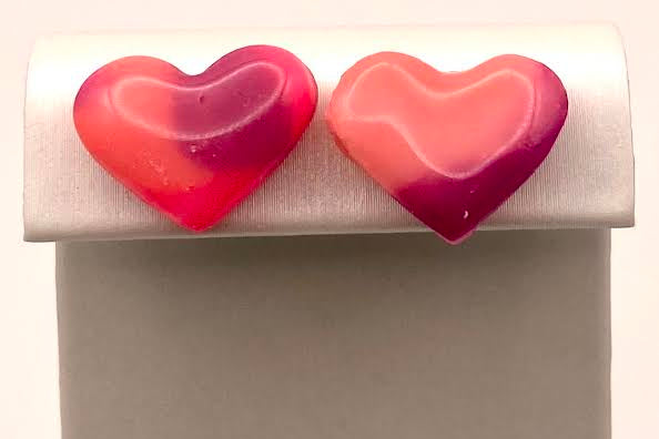 A pair of heart-shaped stud earrings in a glossy marbled pink and red finish, displayed on a white jewelry stand against a light background.