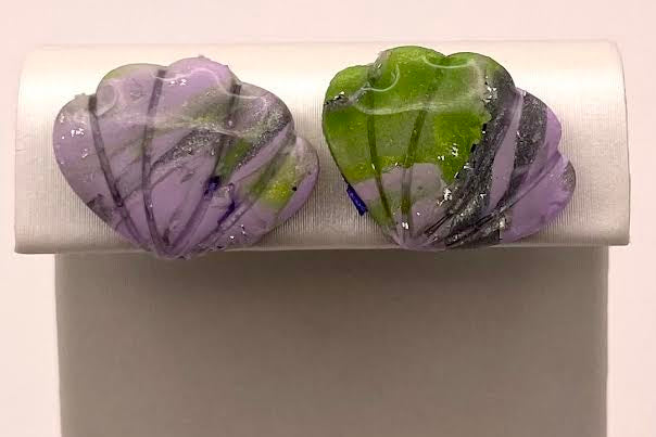 Two handmade glass shell stud earrings displayed on a white jewelry stand. The left earring is lavender purple with black and white marbling, and the right earring is green with black and white marbling.