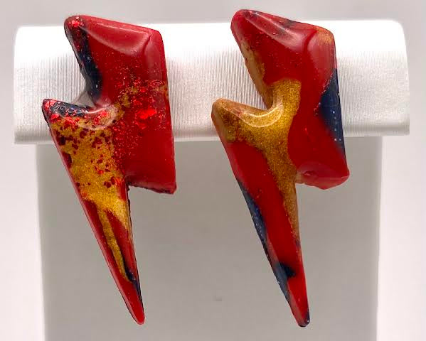 A pair of lightning bolt-shaped earrings with a glossy red finish, accented with gold glitter and dark blue/black speckles, displayed on a white jewelry stand.