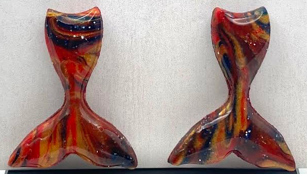 A pair of handmade earrings shaped like stylized mermaid tails, featuring a marbled swirl of red, orange, yellow, and black. They are displayed on a black stand in front of a white textured background card with the brand name 'Phoenix Frost Art' printed in black script.